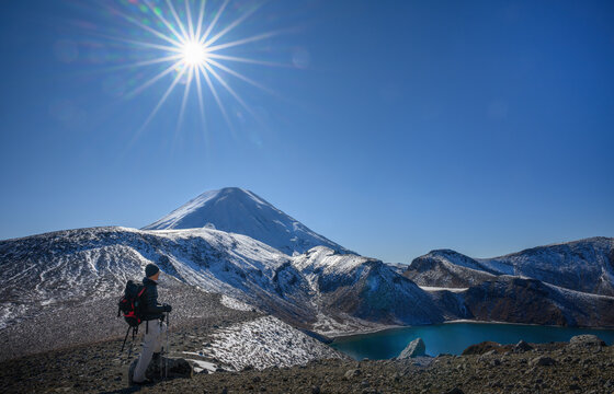 Hiker admiring the Upper Tama Lake. Sun starbursts shining over snow-capped Mount Ngauruhoe. Tongariro National Park. New Zealand.
