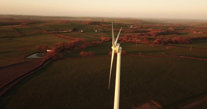 Aerial view of a wind turbine standing in a rural landscape with rolling green fields and hedgerows during golden hour in Devon, England, United Kingdom.
