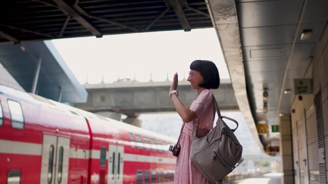 The girl in a pink dress and with a camera stands on the platform of the train station and waves with her hand. goodbye to friends. travel and vacation. creating content about travel. tourist with cam