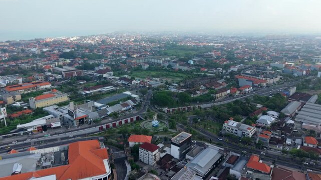 Aerial view of the Statue of Dewa Ruci roundabout surrounded by urban buildings, roads, and lush greenery in Kecamatan Kuta, Bali, Indonesia.