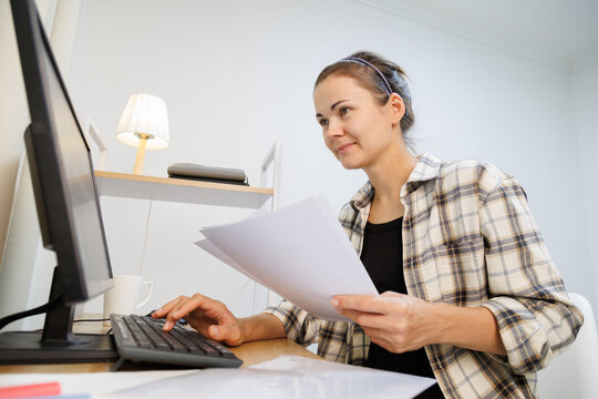 Working with paper documents and computer at a home desk, comparing information, checking records and completing business paperwork.