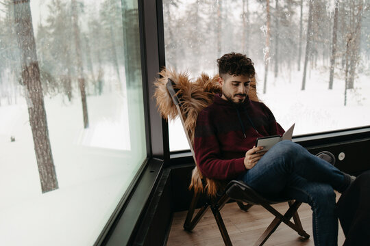 Young man reading in a cozy glass cabin in snowy Lapland