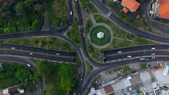 Aerial view of the Statue of Dewa Ruci in the center of a busy roundabout with surrounding roads, lush green trees, and traffic in Kecamatan Kuta, Bali, Indonesia.