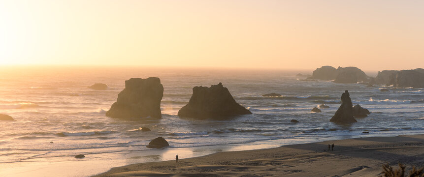 View of the iconic sea stacks at Face Rock State Scenic Viewpoint during a golden sunset with people walking along the sandy beach in Bandon, Oregon, United States.