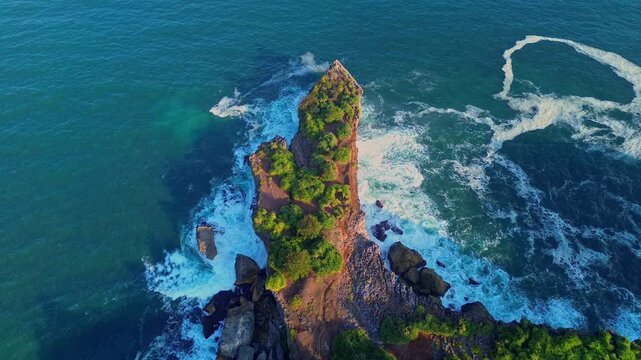 Aerial view of Baron Beach featuring a rocky cliff covered in lush green vegetation surrounded by turquoise ocean waves in Yogyakarta, Special Region of Yogyakarta, Indonesia.