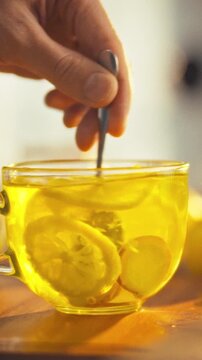 Man's hand stirs tea in clear glass mug, a lemon slice and ginger pieces swirl in the hot water, visible zest floats around, presenting a beverage and food preparation scene in a warm morning setting