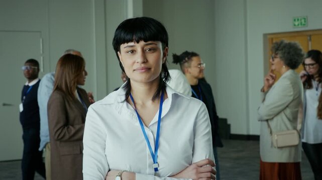 A focused young professional woman with crossed arms stands confidently amidst a bustling business conference, surrounded by diverse colleagues networking and engaging in conversations at the event.