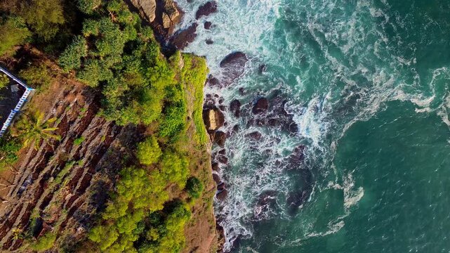 Aerial view of Baron Beach with rugged cliffs, lush green vegetation, and turquoise waves crashing against the rocky shore in Yogyakarta, Special Region of Yogyakarta, Indonesia.