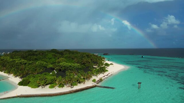 Aerial view of Meedhupparu island featuring white sand beaches, lush palm trees, turquoise lagoon waters, and a vibrant rainbow in the sky, Raa Atoll, Maldives.