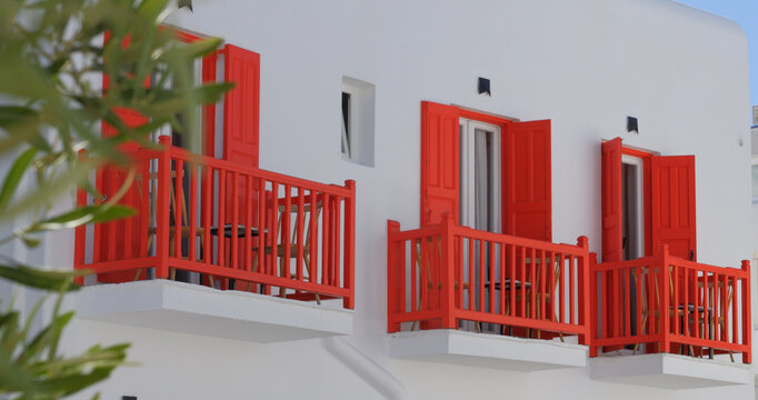 View of a traditional white building facade in Mykonos featuring vibrant red shutters and balconies, with an olive tree branch in the foreground, Mykonos, Greece.
