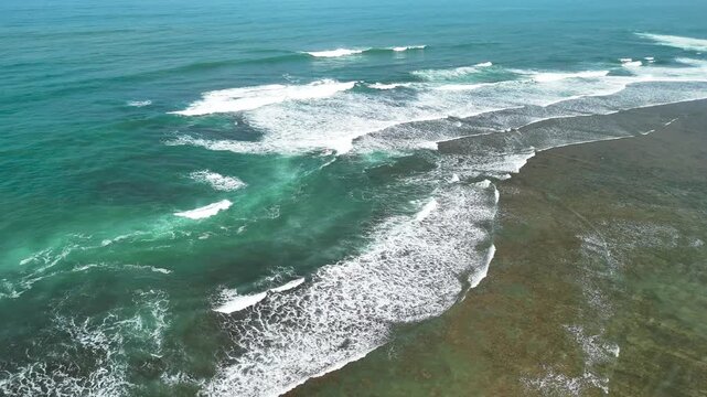 Aerial view of turquoise ocean waves breaking over a shallow coral reef with clear water and coastal scenery in Indonesia.