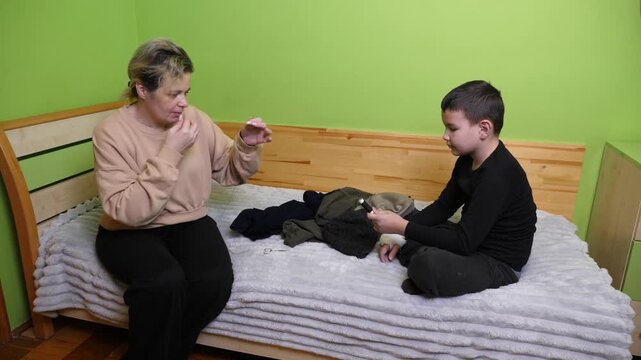 A mother teaches her son to sew and repair clothing, demonstrating thread and needle work during a home activity.