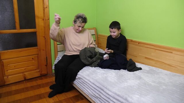 A mother sews clothing with needle and thread while her son watches and helps during a home repair activity.