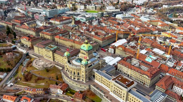 Aerial drone view of the Federal Palace of Switzerland with its copper dome and Swiss flags, the Bundesplatz ice rink, Bundesterrasse and old town rooftops, Bern, in winter.