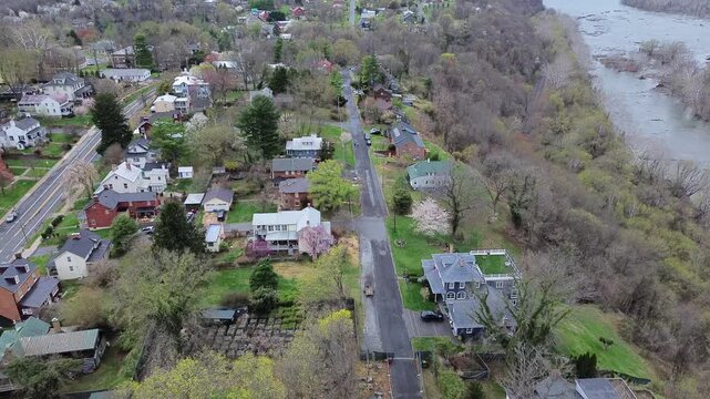 Single lane asphalt roadway hugs steep wooded bluff beside rushing Potomac River shallows. Colorful historic cottages overlook Appalachian mountain valley in Harpers Ferry, West Virginia