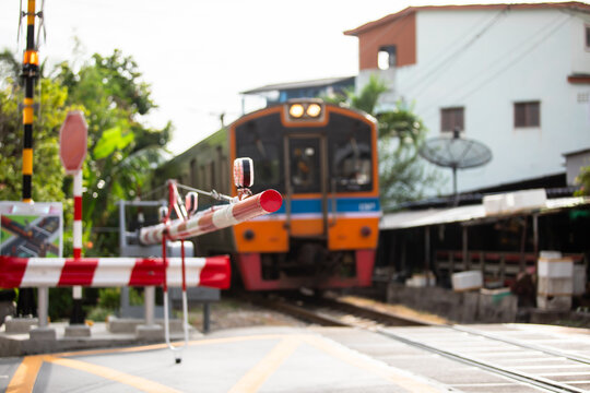 Selective focus to railroad crossing gate with blurry passenger train approaching a closed level crossing safety barrier in a suburban community in Thailand, featuring warning lights, signals.