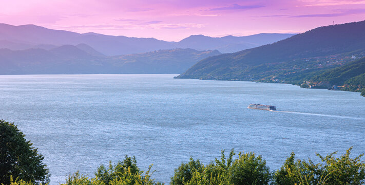 A river cruise ship glides along a wide Danube gorge beneath a vivid pink and purple twilight sky, framed by layered mountain silhouettes. Travel concept.