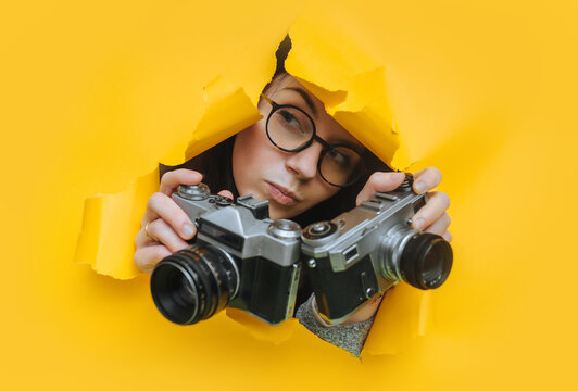 A paparazzi girl in glasses with two rare SLR cameras looks out from her hiding place, watching what is happening. Yellow paper, torn hole. Tabloid press. Looking for an item, stock photo.