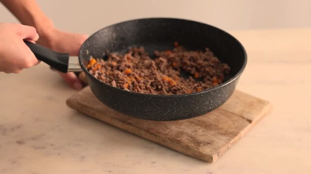 Hand sets rustic wooden board on marble countertop, then lowers nonstick skillet filled with browned ground beef and diced carrots onto it, preparing bolognese sauce base in bright modern kitchen.