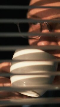 Individual wearing glasses sips from a white cup while looking through partially closed window blinds, warm evening light fills the room, in a modest living area, creating a quiet domestic atmosphere