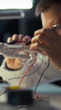 In well-lit engineering lab at night, engineer assembles robotic hand prototype, routing wires and calibrating sensors among scattered tools and monitor screens, advancing robotics development concept