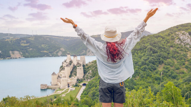 Red-haired woman in hat raises arms joyfully overlooking Golubac Fortress and Danube River