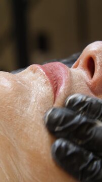 Professional beautician applying cleansing mask on female client's face. Close-up of female face during youth restoration and facial rejuvenation procedure  - Vertical Video