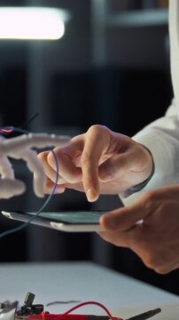 In laboratory setting with precision instruments, researcher in lab coat operates a tablet while assembling a robotic hand, integrating advanced technology and robotics for scientific testing concept