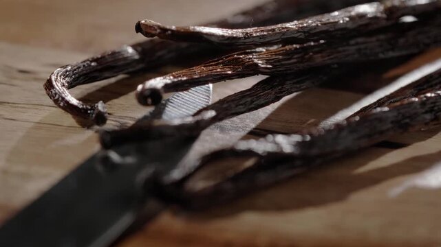 Cinematic macro close-up featuring a forward dolly-in motion toward vanilla bean pods positioned opposite a knife on a wooden cutting board. Shallow depth of field and warm lighting highlight their ri
