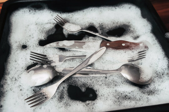 A knife with wooden handle, fork and spoons in the detergent foam on a black oven-tray. Washing dishes concept.