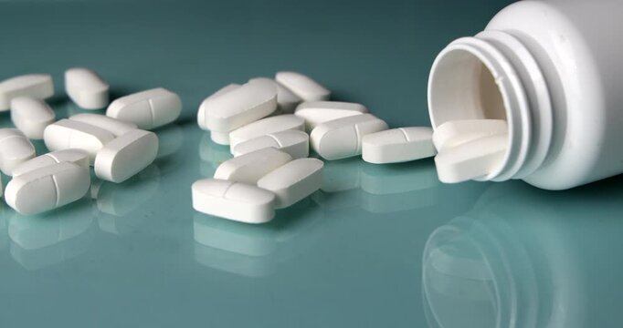 Smooth horizontal camera pan gliding over scattered white medicine pills to reveal a white plastic drug bottle on a light blue glass tabletop, clinical healthcare and pharmacy layout