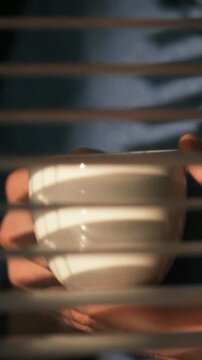 At home in the early morning, a person's hand grips a small white cup while looking through partially closed window blinds, depicts a quiet contemplation as soft light filters into the room