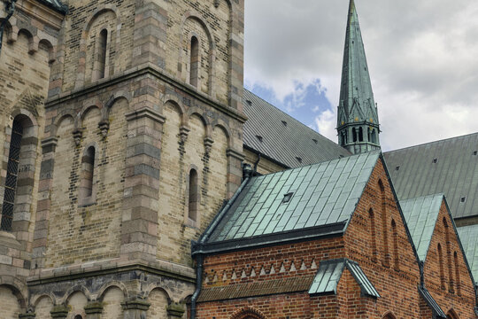 View of the historic Ribe Cathedral featuring intricate Romanesque brickwork, arched windows, and a distinctive green copper spire under a cloudy sky in Ribe, Denmark.