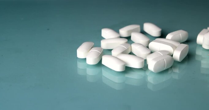 Horizontal slider movement panning across white medical pills and ending on a white pill bottle resting on a light blue glass table, pharmacy drug therapy and healthcare concept