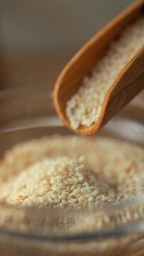 A man's hand pours sesame seeds from a small spice scoop into a clear glass bowl in a kitchen, part of meal preparation, using a culinary ingredient for cooking, within a food preparation process