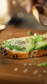 A man prepares his breakfast in the kitchen by delicately sprinkling sesame seeds onto a sandwich filled with avocado and cream cheese, adding texture and nutrients as part of food preparation