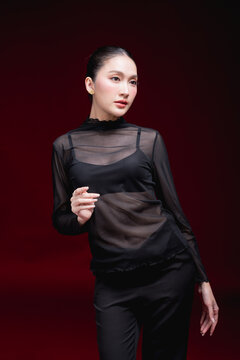 Full-length editorial portrait of a young Asian woman in a sheer black top and pants, touching her hair with a striking gaze against a deep red background.