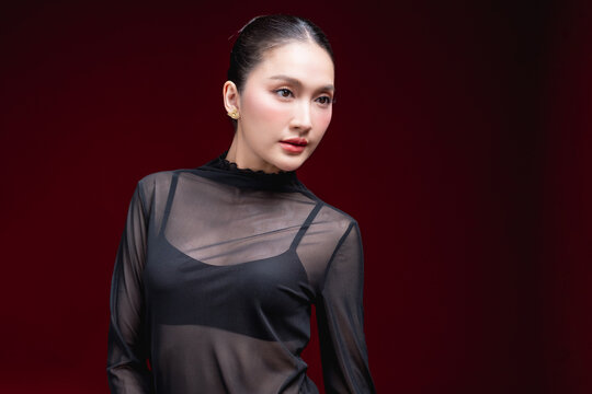 Side-profile beauty portrait of a young Asian woman in a sheer black top looking off to the side with a subtle expression against a deep red background.