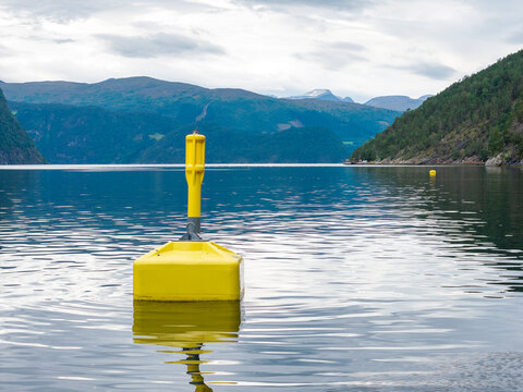 View of a bright yellow navigation buoy floating in the calm waters of a fjord, with lush green mountains under a cloudy sky in Liabygda, More og Romsdal, Norway.