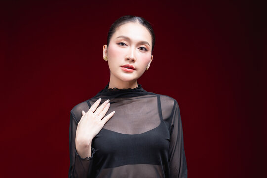 Cheerful close-up beauty portrait of a young Asian woman in a sheer black top laughing warmly with a raised hand near her face against a deep red background.