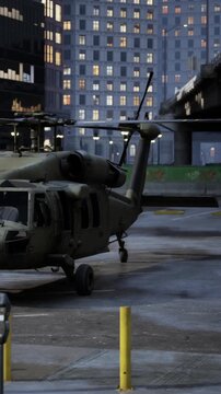 A military helicopter sits on a city street. Buildings surround the area with lights starting to turn on. The scene shows the helicopter at rest during early evening.
