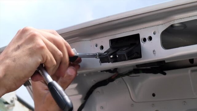 Mechanic using screwdriver to install metal latch mechanism on vehicle trunk lid.