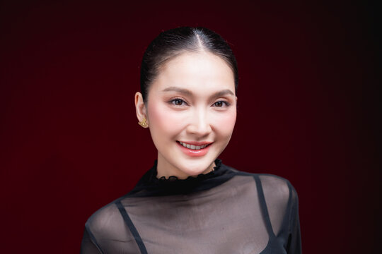 Cheerful close-up beauty portrait of a young Asian woman in a sheer black top laughing warmly with a raised hand near her face against a deep red background.