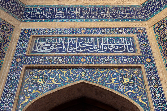 Ornate tilework and Arabic calligraphy above a pointed arch at Kalyan Mosque in Bukhara. Intricate floral and geometric mosaic patterns in blue, turquoise, and gold showcase Islamic decorative arts.