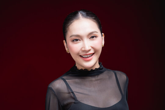 Cheerful close-up beauty portrait of a young Asian woman in a sheer black top laughing warmly with a raised hand near her face against a deep red background.