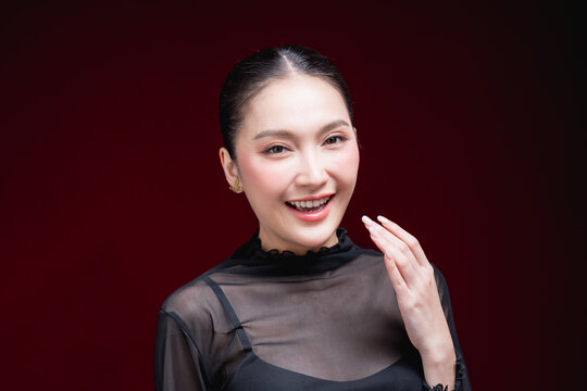 Close-up beauty portrait of a young Asian woman in a sheer black top holding both hands to her head, gazing to the side against a deep red background.