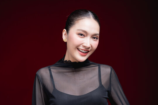 Close-up beauty portrait of a young Asian woman in a sheer black top holding both hands to her head, gazing to the side against a deep red background.