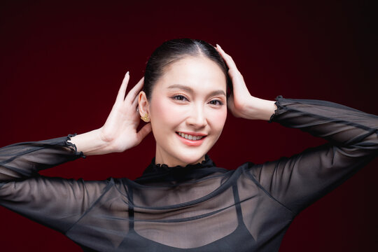 Close-up beauty portrait of a young Asian woman in a sheer black top holding both hands to her head, gazing to the side against a deep red background.