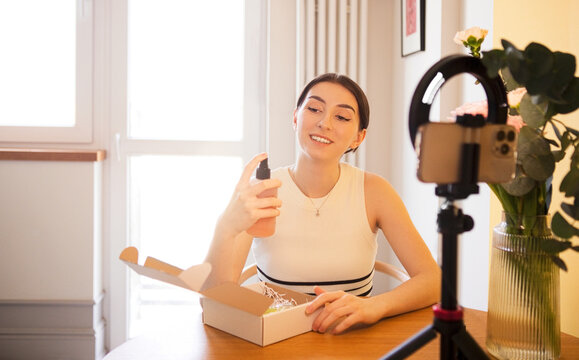 Young woman with brown hair in white top holds a skincare product while sitting at a table with a ring light and flower vase in a bright indoor setting