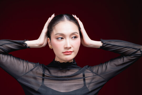 Close-up beauty portrait of a young Asian woman in a sheer black top holding both hands to her head, gazing to the side against a deep red background.